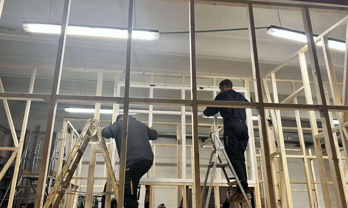 Es ist eine beleuchtete Halle zu sehen in der das Holzrahmengestell eines »Tiny Houses« von zwei Mitarbeitenden mit einem großen Fenster bestückt wird.